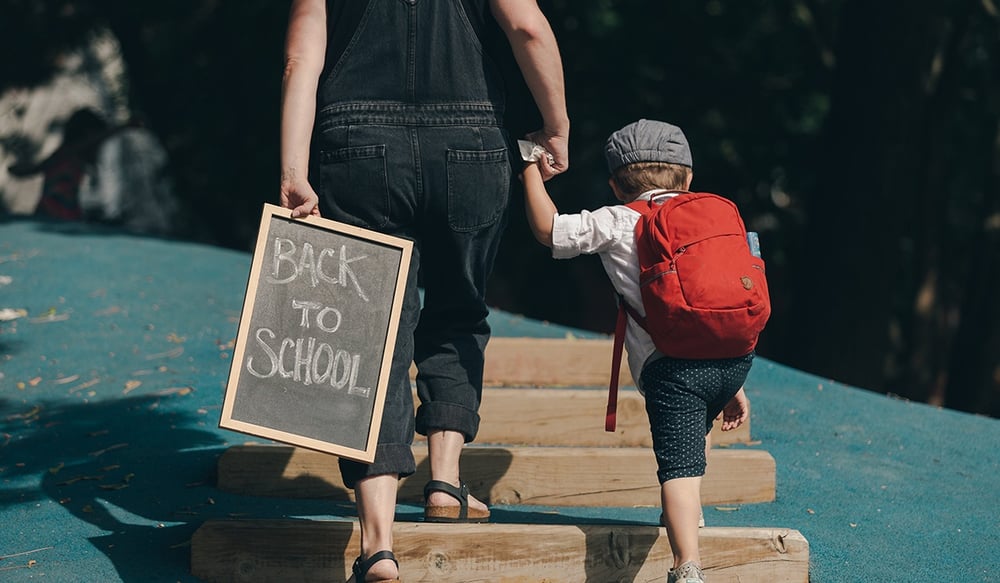 Mom walking young boy to school with sign in hand that reads - Back to School