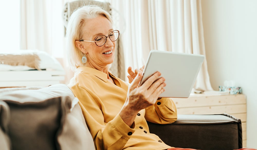 A smiling senior woman using a tablet while sitting on a couch in her home